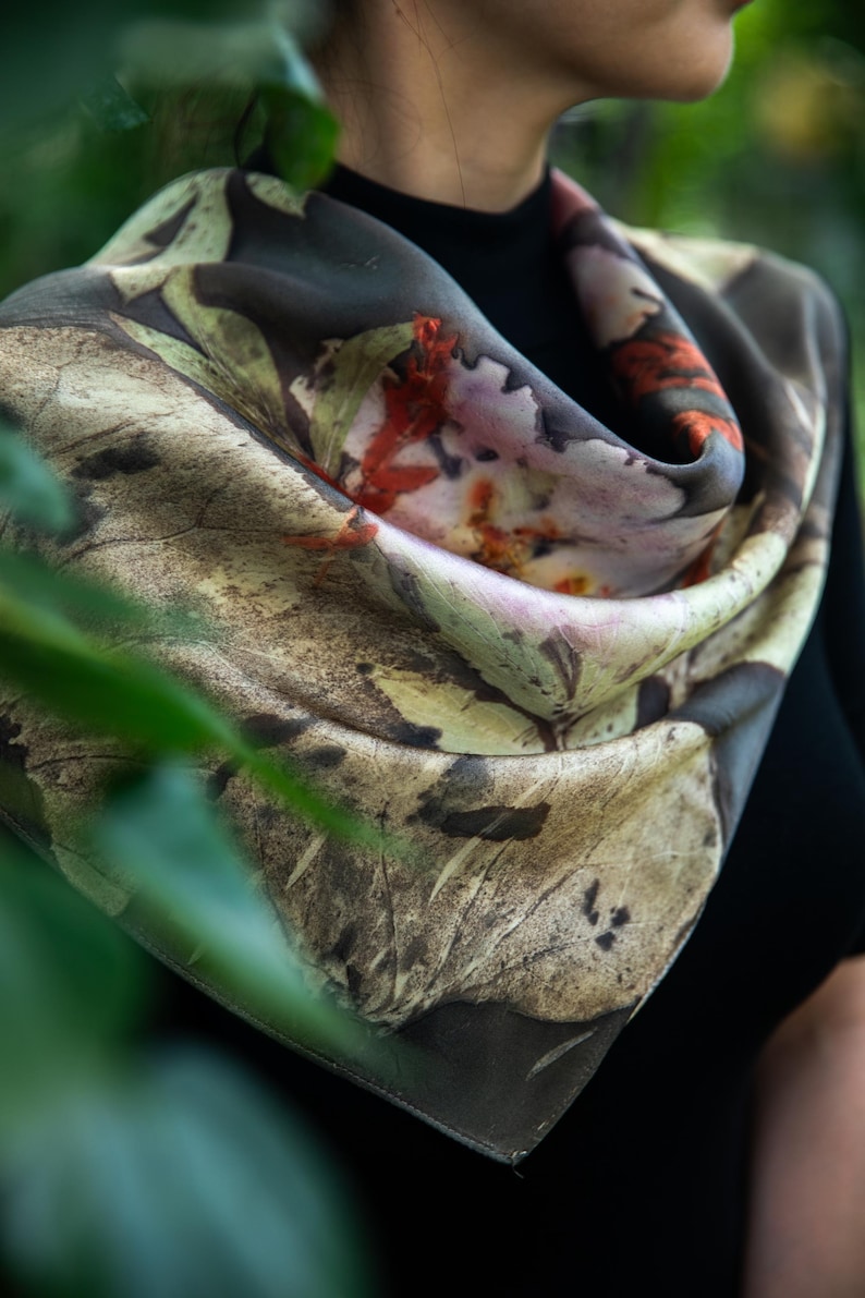 May include: A square scarf featuring a botanical print with earthy tones of brown, beige, and muted green, accented by vibrant orange and deep red leaf patterns. The fabric displays intricate, detailed textures of foliage and organic plant imprints.