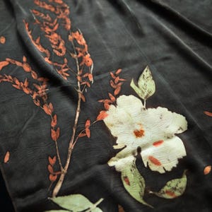 May include: A dark charcoal gray fabric featuring botanical prints of a large white flower with orange accents and a tall, slender branch with small, elongated orange leaves, alongside a single light green leaf at the bottom edge.