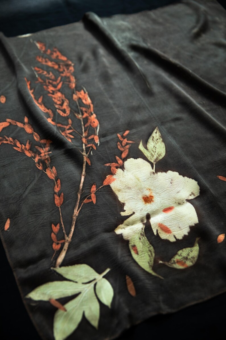 May include: A dark charcoal gray fabric featuring botanical prints of a large white flower with orange accents and a tall, slender branch with small, elongated orange leaves, alongside a single light green leaf at the bottom edge.