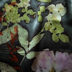 May include: A dark charcoal fabric featuring a botanical print with pressed green leaves, reddish-brown stems, and a large pale pink flower. The natural plant imprints create a detailed, organic pattern across the smooth, slightly reflective textile surface.