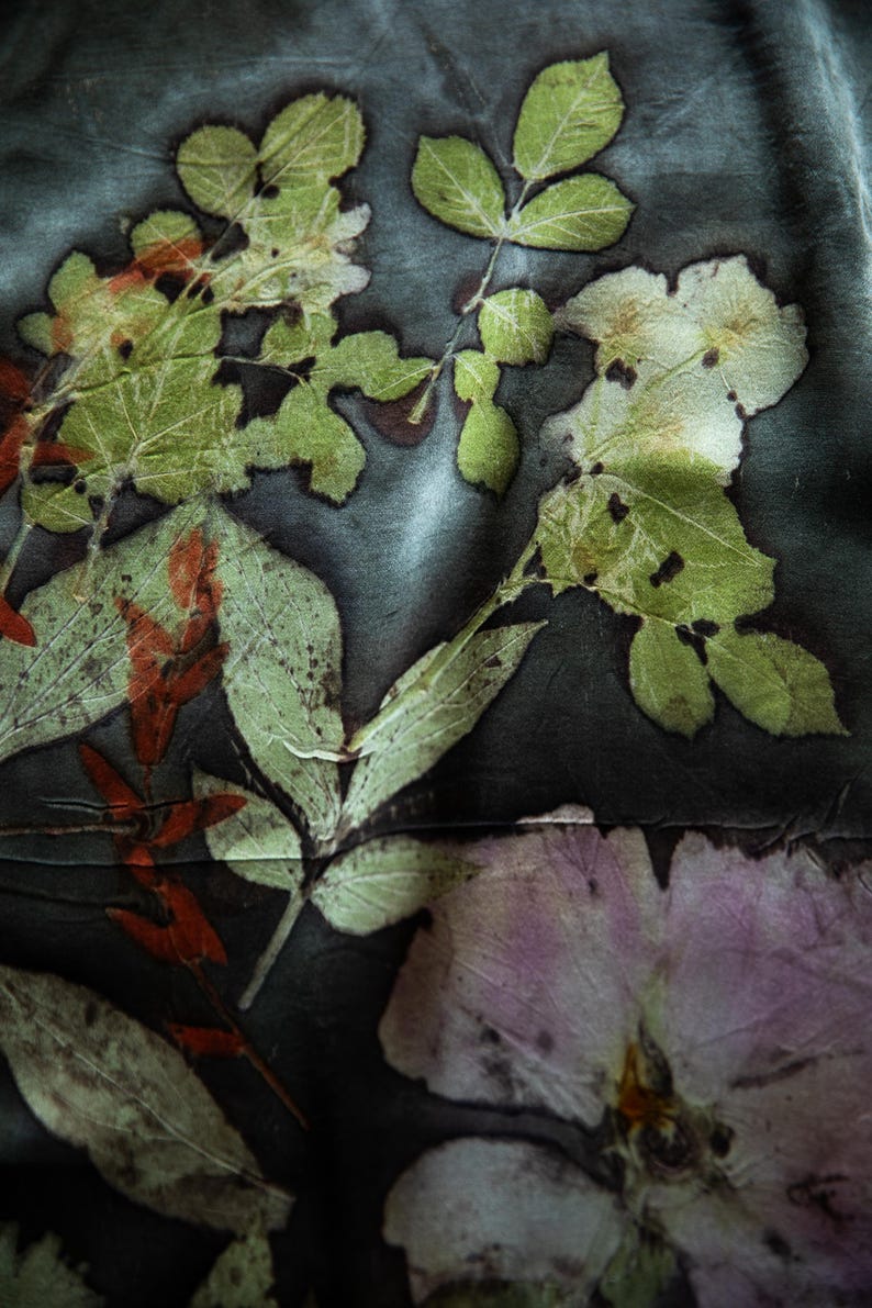 May include: A dark charcoal fabric featuring a botanical print with pressed green leaves, reddish-brown stems, and a large pale pink flower. The natural plant imprints create a detailed, organic pattern across the smooth, slightly reflective textile surface.