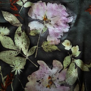 May include: Close-up of a textile with a botanical print. The design features large, pale pink flowers and green leaves with a dark background. The leaves and flowers have a textured, pressed appearance, creating a natural, artistic aesthetic.
