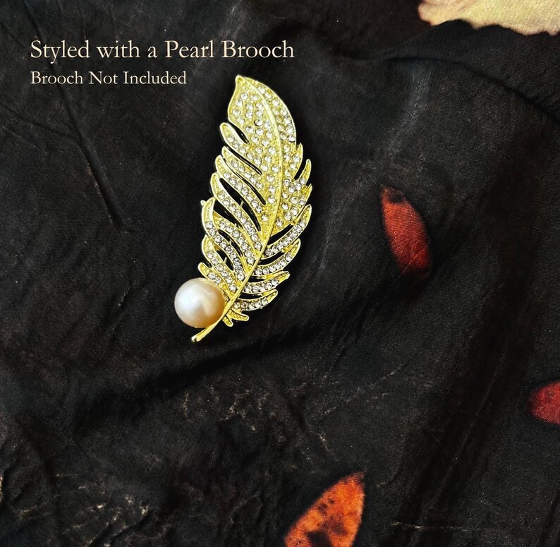 May include: A gold-toned feather-shaped brooch encrusted with clear rhinestones and featuring a single round white pearl at the base. The text on the image reads: Styled with a Pearl Brooch. Brooch Not Included.