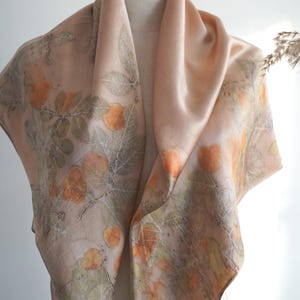 May include: A square silk scarf in a soft peach hue featuring a botanical print of orange fruit and green leaves. The design displays detailed leaf veins and rounded fruit shapes scattered across the fabric, creating a natural, earthy pattern.
