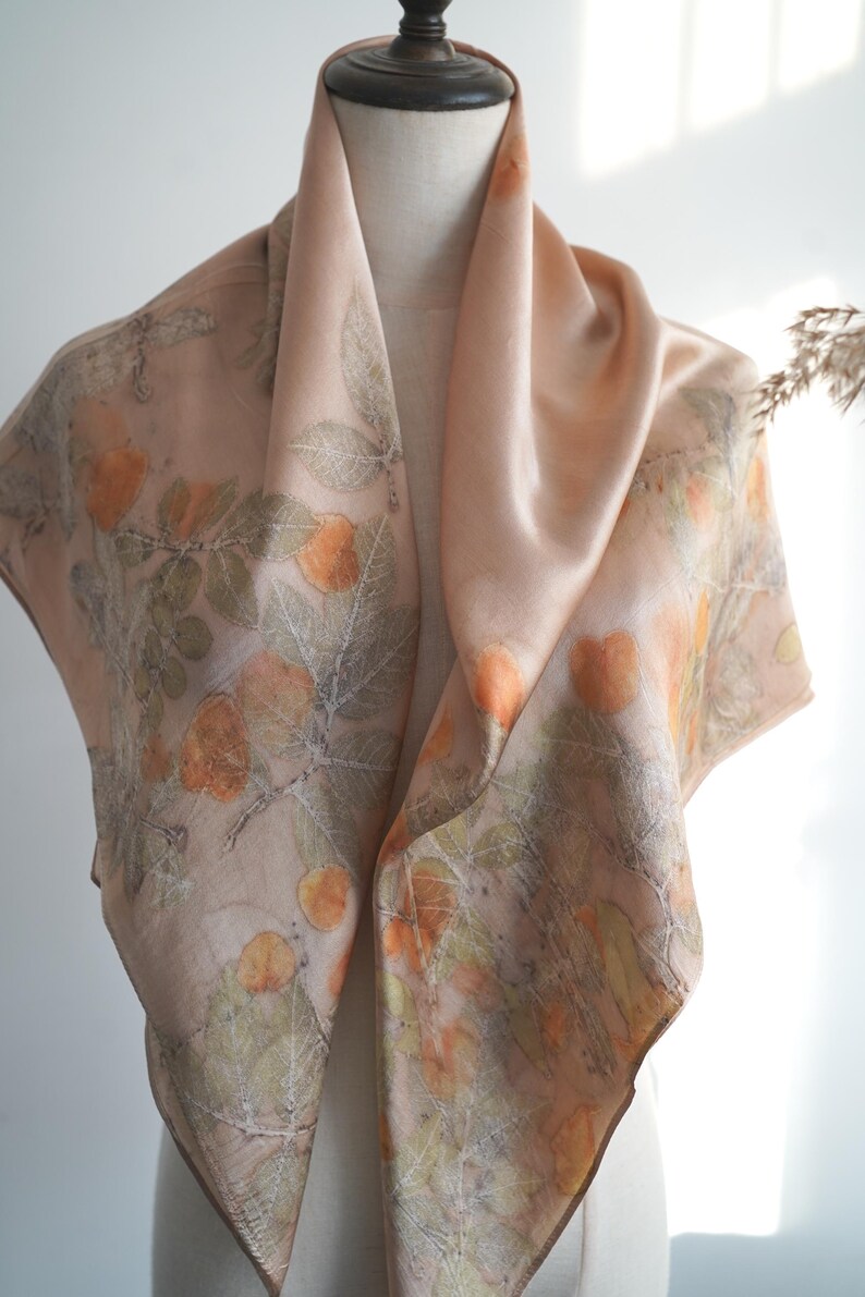 May include: A square silk scarf in a soft peach hue featuring a botanical print of orange fruit and green leaves. The design displays detailed leaf veins and rounded fruit shapes scattered across the fabric, creating a natural, earthy pattern.
