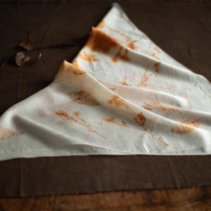 May include: A triangular white scarf featuring botanical prints in shades of orange and brown. The fabric displays delicate, detailed imprints of leaves and stems scattered across the surface, creating a natural, earthy pattern on the smooth material.