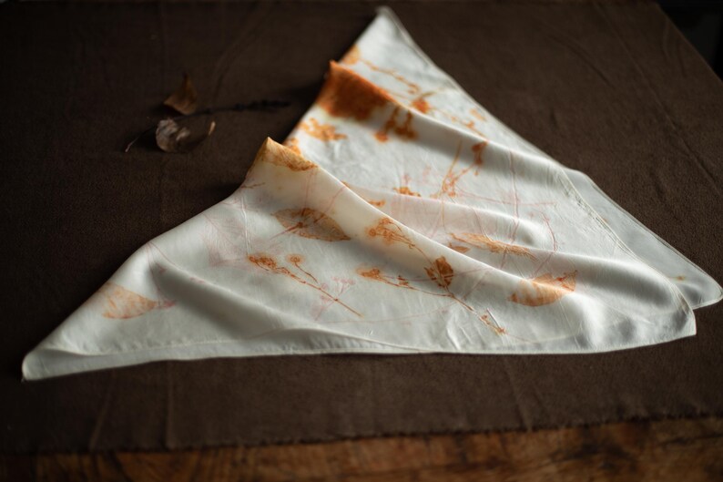 May include: A triangular white scarf featuring botanical prints in shades of orange and brown. The fabric displays delicate, detailed imprints of leaves and stems scattered across the surface, creating a natural, earthy pattern on the smooth material.