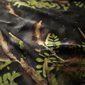 May include: A dark black fabric featuring a botanical print of green and golden-brown fern leaves and foliage. The material has a soft, draped texture with visible folds and creases that create depth across the surface of the patterned textile.