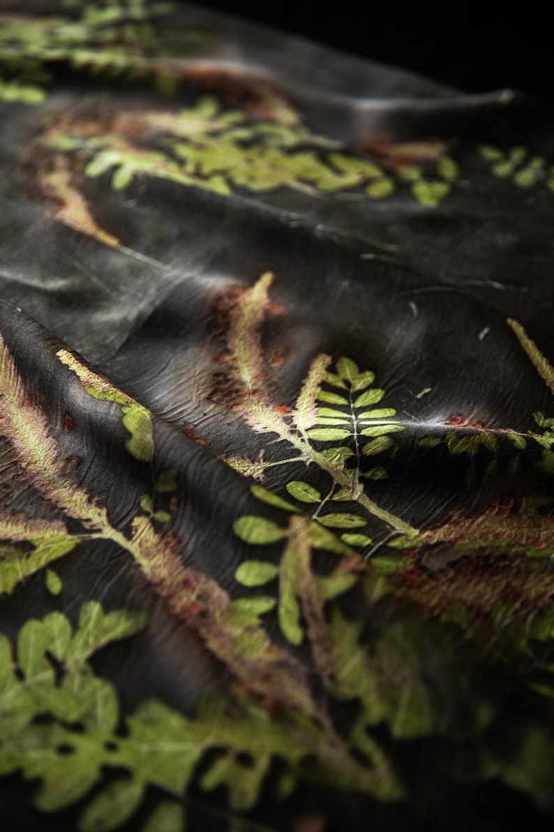 May include: A dark black fabric featuring a botanical print of green and golden-brown fern leaves and foliage. The material has a soft, draped texture with visible folds and creases that create depth across the surface of the patterned textile.