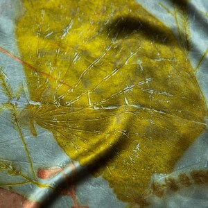 May include: A light gray fabric featuring a large, prominent mustard yellow leaf print with visible veins. The textile displays additional smaller, delicate botanical imprints in muted orange and brown tones, creating a layered, organic pattern across the surface.