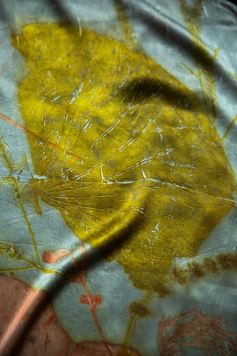 May include: A light gray fabric featuring a large, prominent mustard yellow leaf print with visible veins. The textile displays additional smaller, delicate botanical imprints in muted orange and brown tones, creating a layered, organic pattern across the surface.
