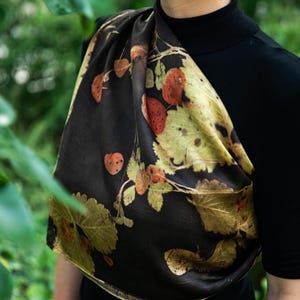 May include: A dark brown scarf featuring a botanical print of various leaves in shades of green, yellow, and burnt orange. The scarf is draped over a black mock neck top, showcasing a smooth, satin-like texture with a repeating natural foliage pattern.