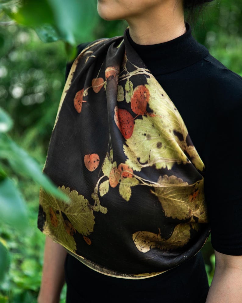May include: A dark brown scarf featuring a botanical print of various leaves in shades of green, yellow, and burnt orange. The scarf is draped over a black mock neck top, showcasing a smooth, satin-like texture with a repeating natural foliage pattern.