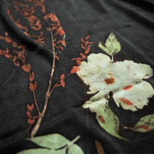 May include: A dark charcoal fabric featuring botanical prints of a large pale green flower and a slender branch with small orange leaves. The textile displays a natural, organic pattern with visible plant textures and earthy color tones throughout.