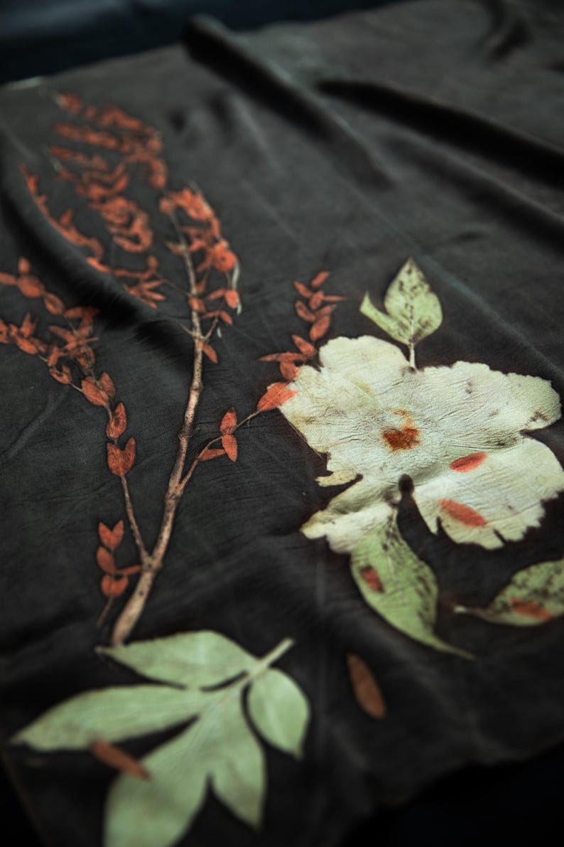 May include: A dark charcoal fabric featuring botanical prints of a large pale green flower and a slender branch with small orange leaves. The textile displays a natural, organic pattern with visible plant textures and earthy color tones throughout.