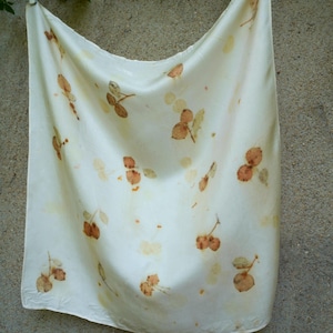 May include: A square off-white fabric scarf featuring a scattered botanical print of small, brown, leaf-like shapes and stems. The fabric has a smooth, slightly reflective surface and is draped in a soft, flowing manner with visible folds.