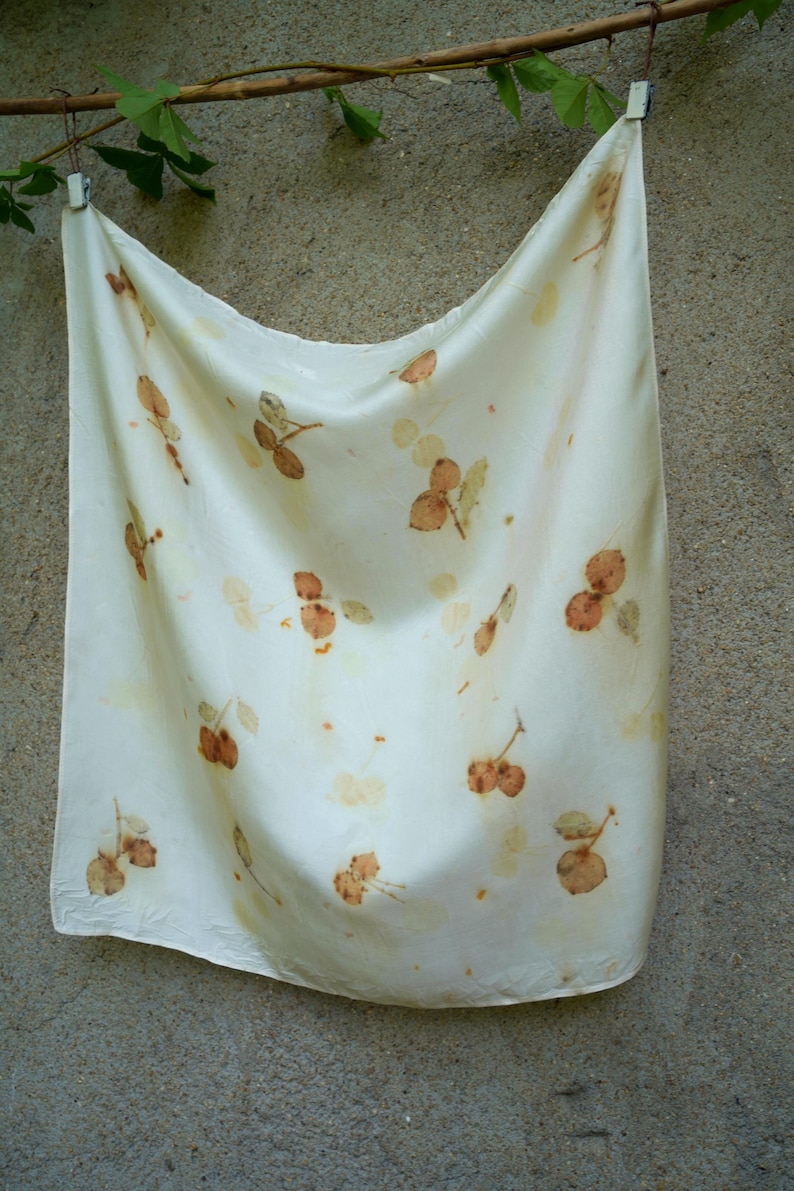 May include: A square off-white fabric scarf featuring a scattered botanical print of small, brown, leaf-like shapes and stems. The fabric has a smooth, slightly reflective surface and is draped in a soft, flowing manner with visible folds.