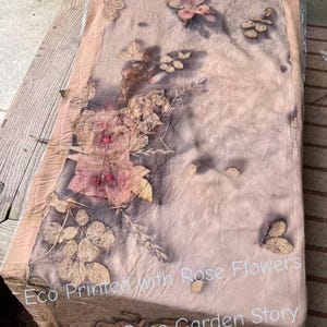 May include: A rectangular fabric runner featuring botanical eco-prints of rose flowers and leaves in shades of brown, tan, and muted pink. The text "Eco Printed with Rose Flowers Telling a Rose Garden Story" is printed in white across the lower section.