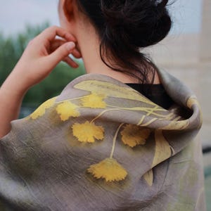 May include: A lightweight scarf featuring a muted taupe and gray base color with a botanical print of yellow flowers and stems. The fabric has a soft, slightly wrinkled texture and is draped over the shoulders, displaying a floral pattern.