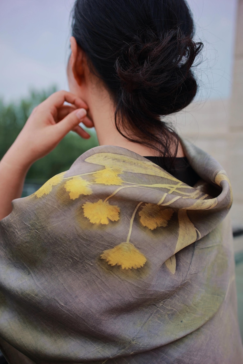 May include: A lightweight scarf featuring a muted taupe and gray base color with a botanical print of yellow flowers and stems. The fabric has a soft, slightly wrinkled texture and is draped over the shoulders, displaying a floral pattern.