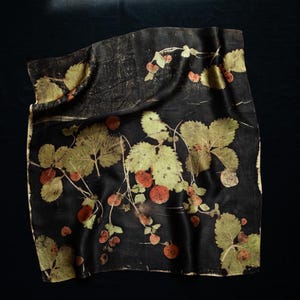 May include: A square textile featuring a dark black background with a botanical print of light green strawberry leaves and small reddish-orange berries. The fabric displays a natural, organic pattern of foliage and fruit scattered across the surface.