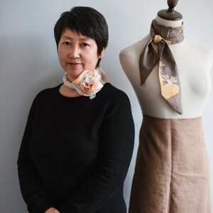 May include: A white silk scarf with peach-colored floral accents tied in a rose shape around the neck, and a brown silk scarf with a yellow botanical print tied around the neck of a dress form wearing a tan skirt.