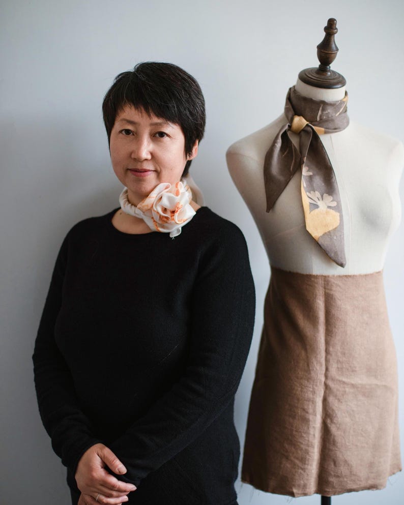 May include: A white silk scarf with peach-colored floral accents tied in a rose shape around the neck, and a brown silk scarf with a yellow botanical print tied around the neck of a dress form wearing a tan skirt.