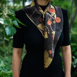 May include: A square scarf featuring a dark brown base with a botanical print of orange strawberries and green leaves. The scarf is tied around the neck, displaying a repeating pattern of fruit and foliage across the fabric surface.