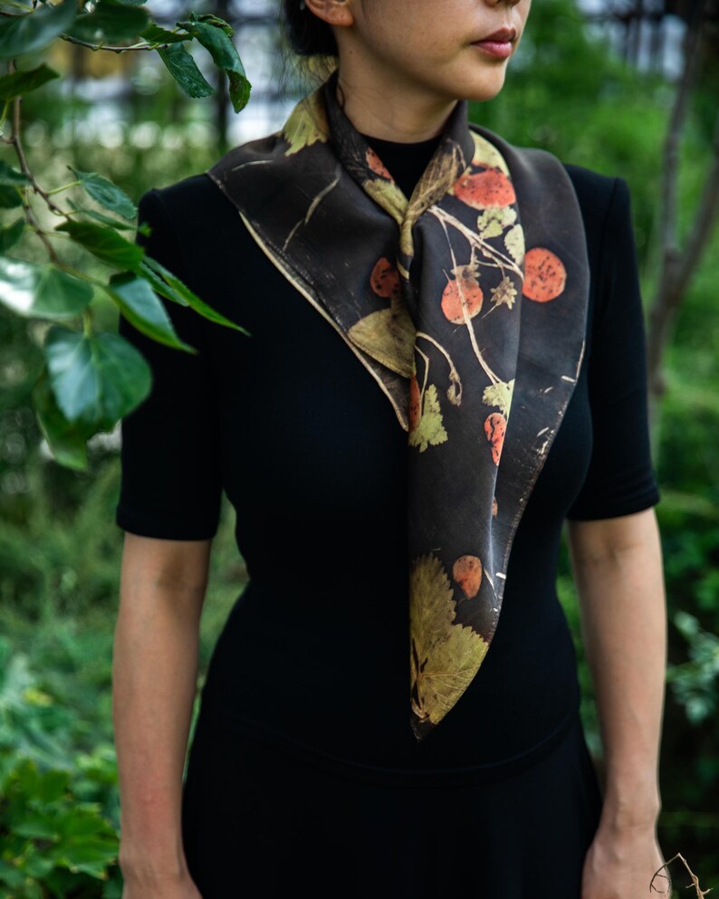May include: A square scarf featuring a dark brown base with a botanical print of orange strawberries and green leaves. The scarf is tied around the neck, displaying a repeating pattern of fruit and foliage across the fabric surface.