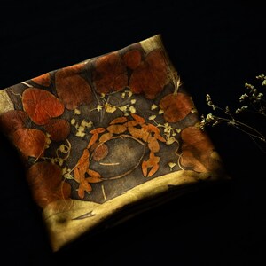 May include: A square textile piece featuring a dark gray base with botanical imprints of reddish-brown rounded leaves and small yellow flowers arranged in a circular pattern, accented by a golden-yellow border and a small sprig of dried flowers.