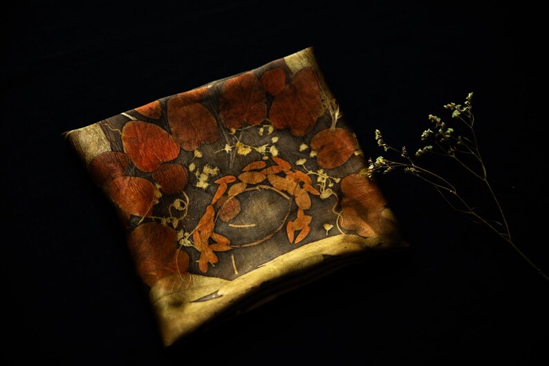 May include: A square textile piece featuring a dark gray base with botanical imprints of reddish-brown rounded leaves and small yellow flowers arranged in a circular pattern, accented by a golden-yellow border and a small sprig of dried flowers.