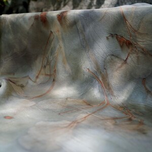 May include: A piece of fabric featuring a botanical print with muted gray and green tones. The textile displays intricate, reddish-brown leaf and branch imprints scattered across the surface, creating a natural, organic pattern with soft folds.