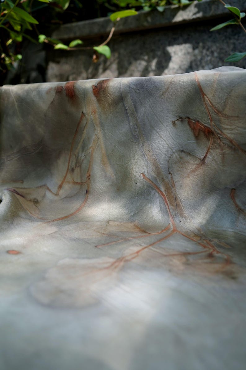 May include: A piece of fabric featuring a botanical print with muted gray and green tones. The textile displays intricate, reddish-brown leaf and branch imprints scattered across the surface, creating a natural, organic pattern with soft folds.