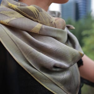 May include: A square scarf featuring a muted taupe and olive green base color with a botanical leaf print pattern in shades of pale yellow and brown. The fabric has a soft, draped texture with finished edges, wrapped loosely around the neck area.