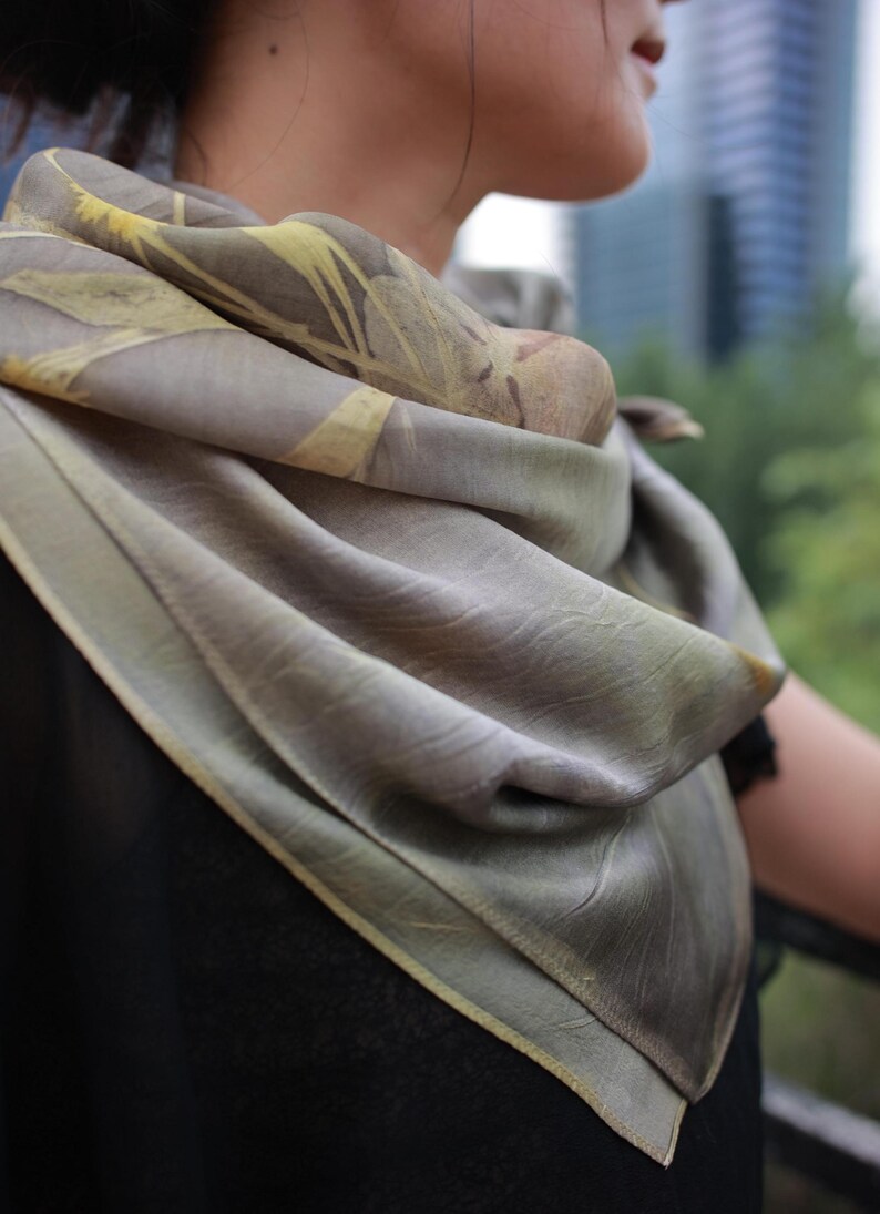 May include: A square scarf featuring a muted taupe and olive green base color with a botanical leaf print pattern in shades of pale yellow and brown. The fabric has a soft, draped texture with finished edges, wrapped loosely around the neck area.