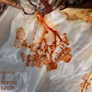May include: A white fabric featuring botanical prints in shades of rust and orange, created through a steaming process. The textile displays detailed leaf and floral patterns transferred onto the surface. Text on the image reads: After steaming, Unwrapping the bundle.