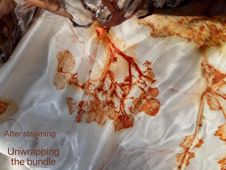 May include: A white fabric featuring botanical prints in shades of rust and orange, created through a steaming process. The textile displays detailed leaf and floral patterns transferred onto the surface. Text on the image reads: After steaming, Unwrapping the bundle.