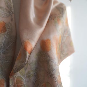May include: A soft, light peach-coloured scarf featuring a botanical print of orange fruit and green leaves. The fabric has a smooth, satin-like sheen and drapes in loose folds, displaying a repeating pattern of natural elements across the surface.