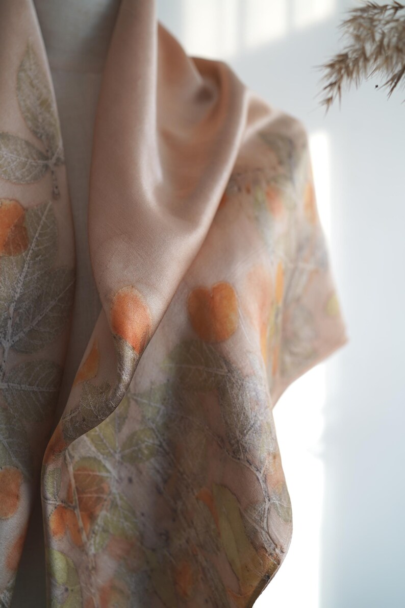 May include: A soft, light peach-coloured scarf featuring a botanical print of orange fruit and green leaves. The fabric has a smooth, satin-like sheen and drapes in loose folds, displaying a repeating pattern of natural elements across the surface.