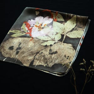 May include: A square silk scarf featuring a dark background with a botanical print of a pale pink flower, green leaves, and a large textured brown leaf. The design includes subtle red accents and a smooth, folded fabric texture throughout the piece.