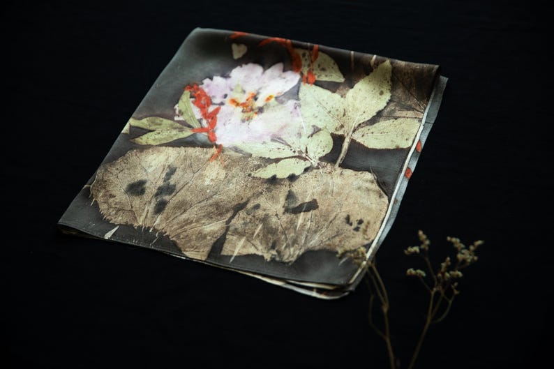 May include: A square silk scarf featuring a dark background with a botanical print of a pale pink flower, green leaves, and a large textured brown leaf. The design includes subtle red accents and a smooth, folded fabric texture throughout the piece.
