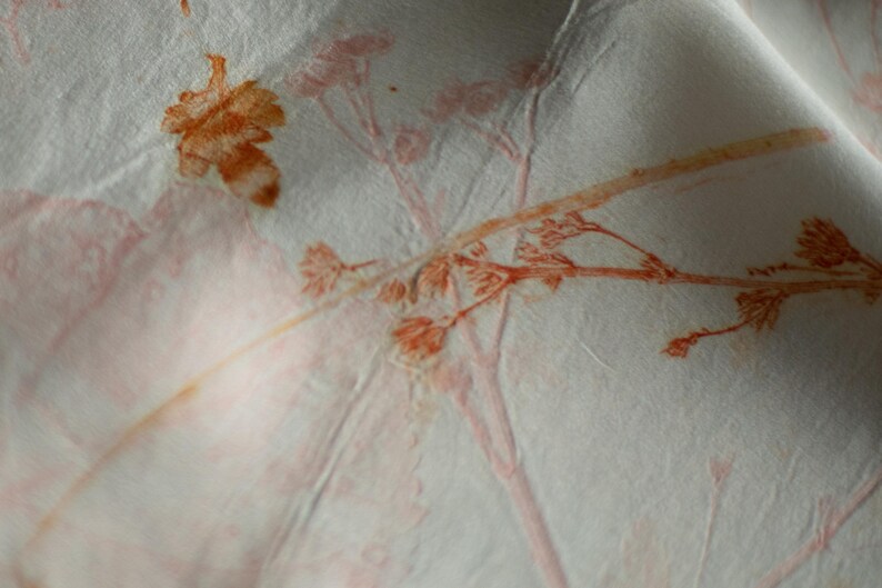 May include: A white fabric surface featuring a botanical print with orange and reddish-brown plant silhouettes. The material has a visible, slightly crinkled texture, and the delicate, detailed imprints of stems, leaves, and small flowers are scattered across the light-colored textile.