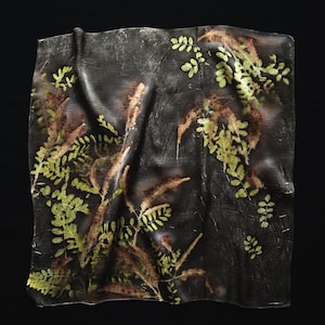 May include: A square fabric piece featuring a dark charcoal gray background with a botanical print of green fern-like leaves and elongated brown textured plant stems. The material has a soft, draped appearance with subtle folds and creases.