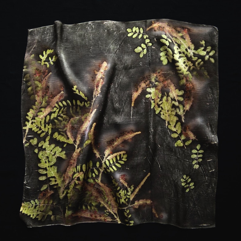 May include: A square fabric piece featuring a dark charcoal gray background with a botanical print of green fern-like leaves and elongated brown textured plant stems. The material has a soft, draped appearance with subtle folds and creases.
