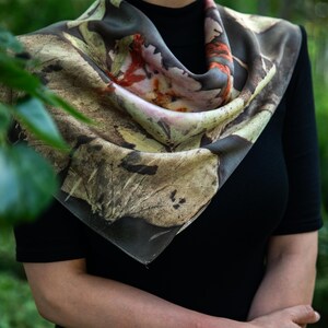 May include: A square silk scarf featuring a botanical print with earthy tones of brown, tan, and muted green, accented by vibrant orange and soft pink floral patterns. The scarf is draped around the neck, displaying a detailed leaf motif.