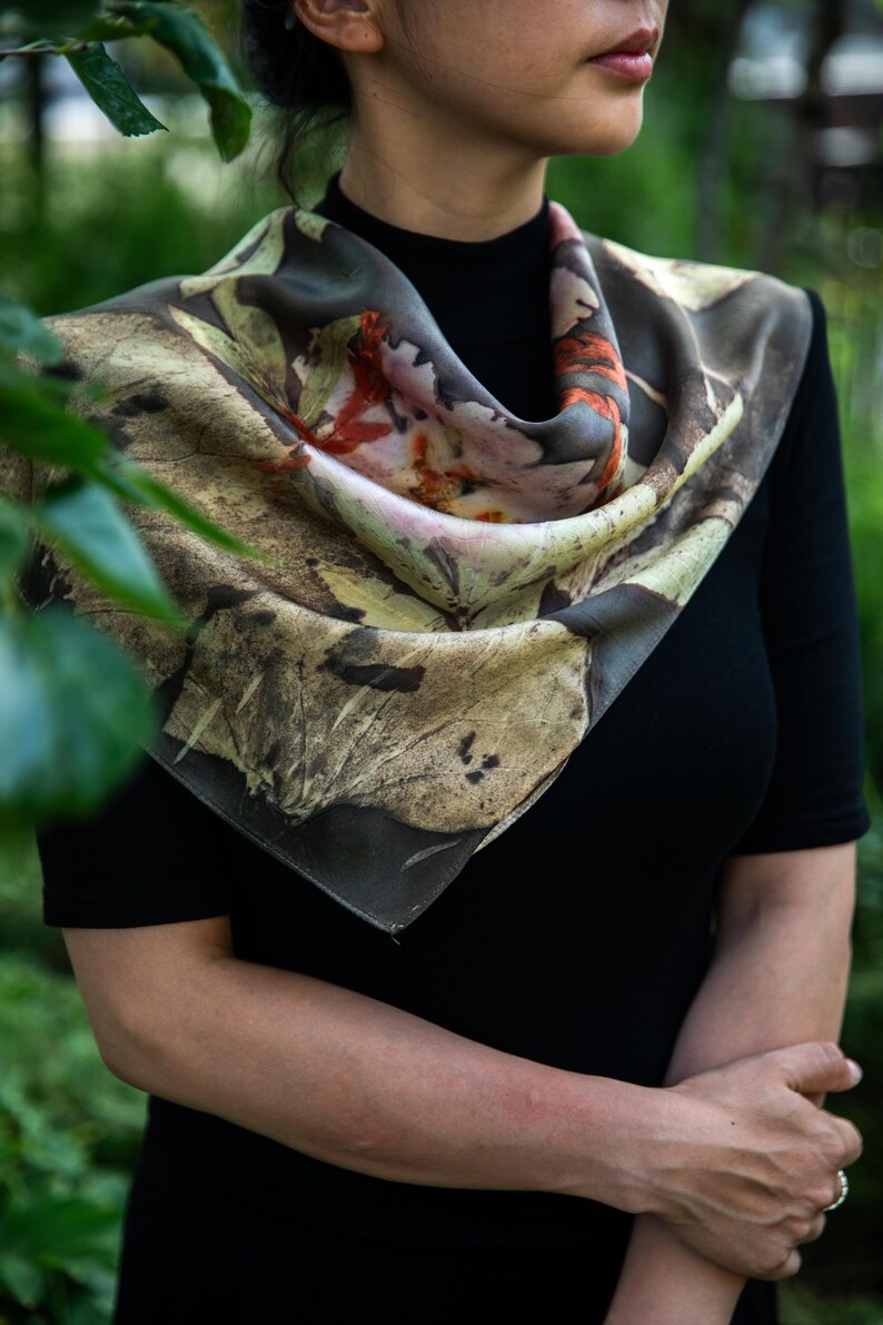 May include: A square silk scarf featuring a botanical print with earthy tones of brown, tan, and muted green, accented by vibrant orange and soft pink floral patterns. The scarf is draped around the neck, displaying a detailed leaf motif.