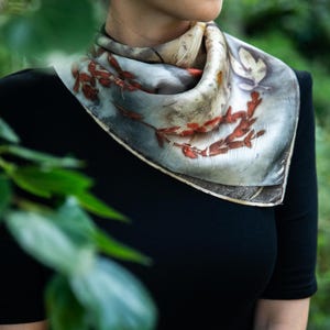 May include: A square silk scarf featuring a botanical print with reddish-brown leaf and branch patterns on a muted grey and cream background. The scarf is folded and tied around the neck, displaying a naturalistic design with visible plant imprints.