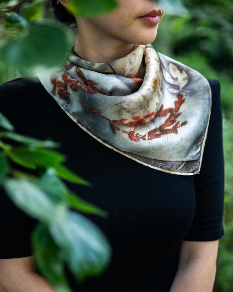 May include: A square silk scarf featuring a botanical print with reddish-brown leaf and branch patterns on a muted grey and cream background. The scarf is folded and tied around the neck, displaying a naturalistic design with visible plant imprints.