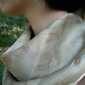 May include: A soft, sage green scarf featuring subtle, organic brown leaf-like imprints and vein patterns across the fabric. The scarf is draped loosely around the neck, showcasing a smooth, matte texture with muted earthy tones and natural markings.