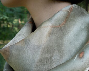 Puede incluir: Una bufanda de color verde salvia suave que presenta sutiles impresiones orgánicas de hojas marrones y patrones de venas en la tela. La bufanda está drapeada holgadamente alrededor del cuello, mostrando una textura suave y mate con tonos tierra.